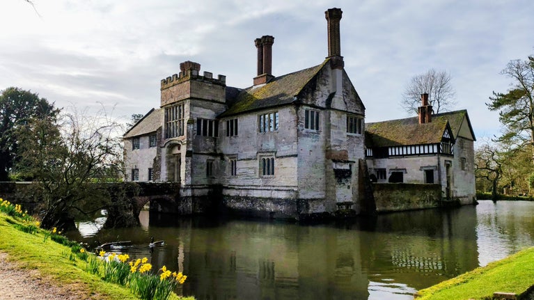 The south-west corner of the house at Baddesley Clinton, Warwickshire, seen across the moat with spring daffodils at the edge